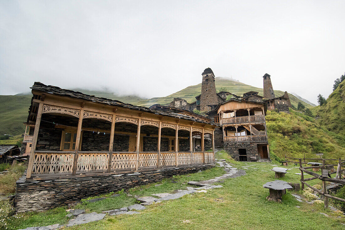 Traditional wooden balconies on the stone houses with medieval watch towers in the background at the mountainside village of Dartlo and the Kvavlo Tower on the mountaintop in the distance in the Tusheti National Park; Dartlo, Kakheti, Georgia
