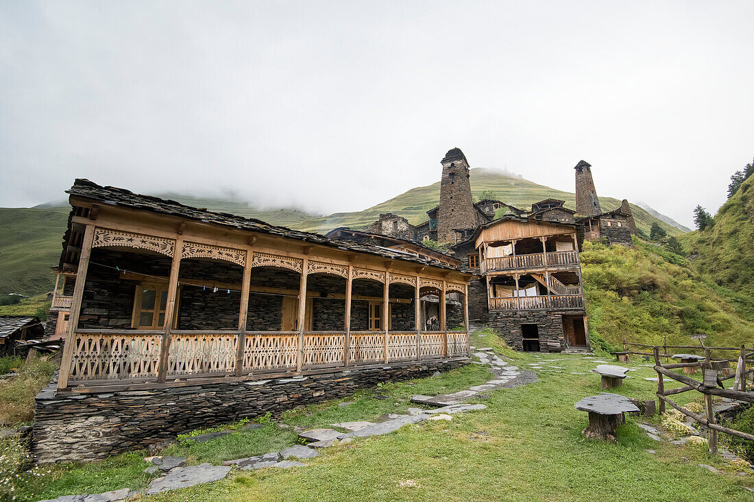 Traditionelle Holzbalkone an den Steinhäusern mit mittelalterlichen Wachtürmen im Hintergrund im Bergdorf Dartlo und dem Kvavlo-Turm auf dem Berggipfel in der Ferne im Tuscheti-Nationalpark; Dartlo, Kachetien, Georgien