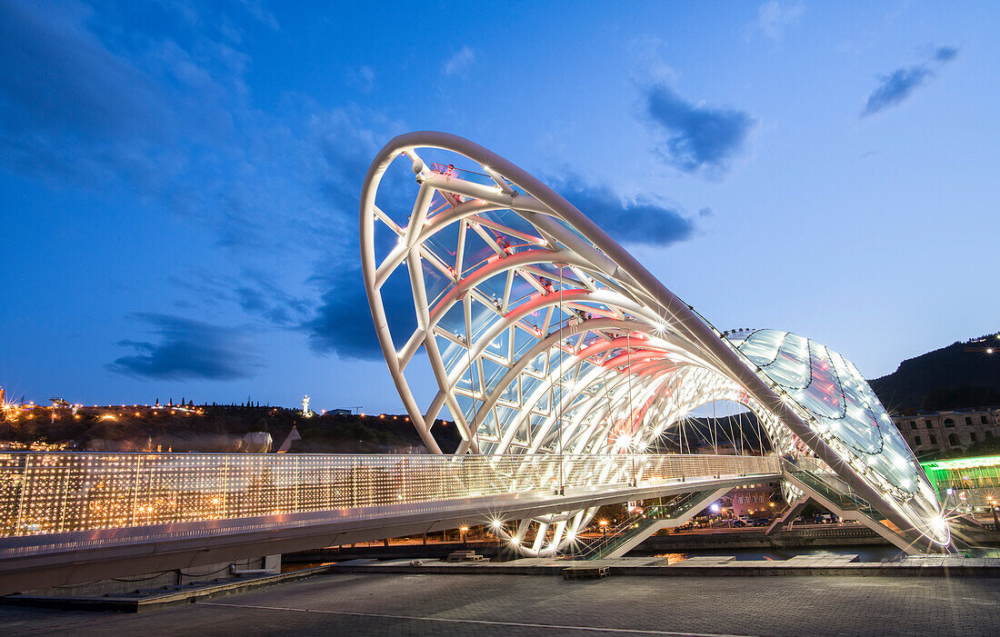 The bow-shaped Bridge of Peace pedestrian bridge, illuminated at dusk spanning the Mtkvari (Kura) River connecting Rike Park and Old Tbilisi; Tbilisi, Georgia