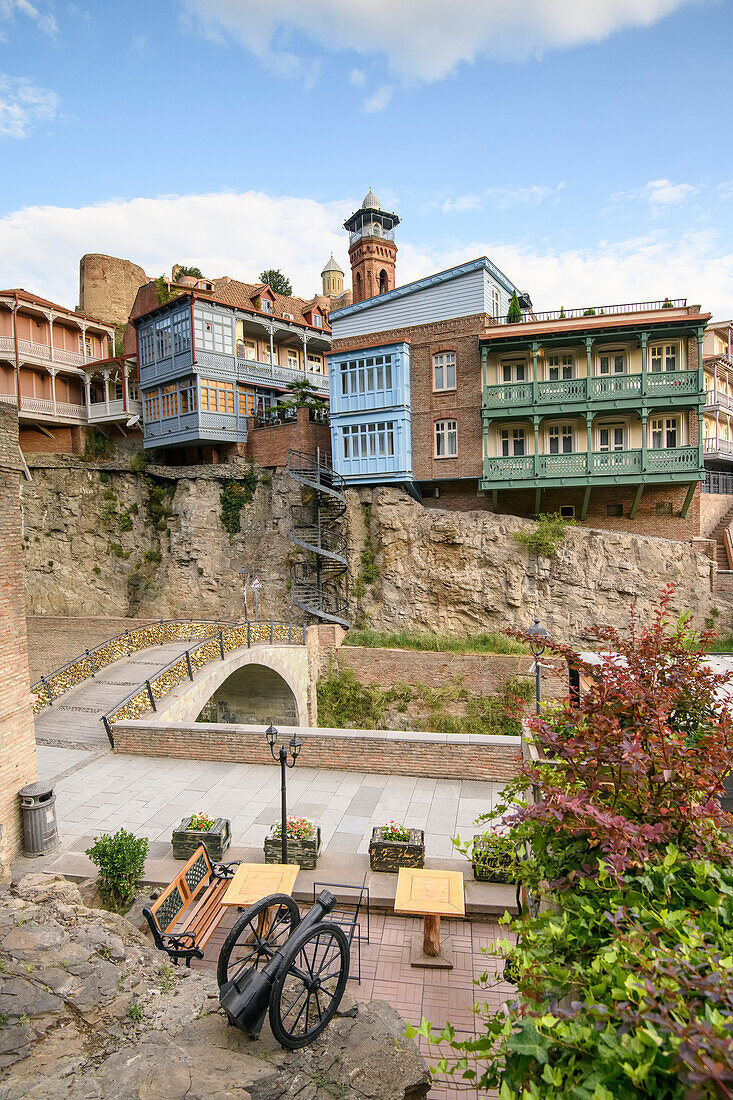 A sitting area with an old cannon next to the small arch bridge crossing the Tsavkisi-Tskali River with love locks attached to its railings in front of the old buildings on the cliffs in Legvtakhevi, part of the historical neighborhood of Abanotubani in the Old Town with the minaret of the Tbilisi Mosque in the background; Tbilisi, Georgia