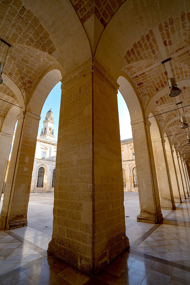 View of the bell tower of the Lecce Cathedral through columns under the portico in front of buildings surrounding the Piazza del Duomo in Lecce; Lecce, Puglia, Italy