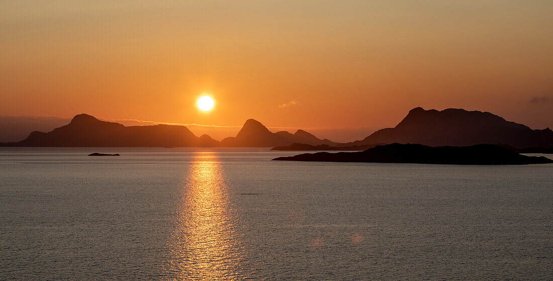 Mitternachtssonne leuchtet hell über den Berggipfeln und spiegelt sich im Nordatlantik; Lofoten, Polarkreis, Norwegen.