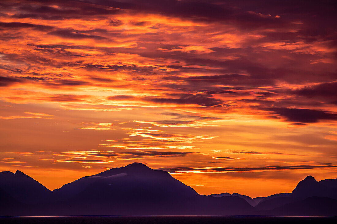 Dramatic cloud formations in the sky, illuminated by the Midnight Sun over the silhouetted mountain peaks in the North Atlantic; Lofoten, Arctic Circle, Norway