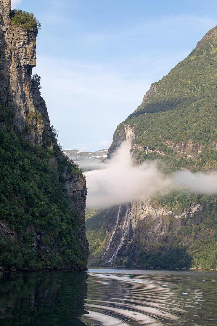 Nahaufnahme der bewaldeten Klippen mit dunstigen Wolken, die Kurve der Wasserstraße, um die spektakuläre Aussicht auf den Sieben-Schwestern-Wasserfall zu sehen, während der Fahrt durch den 15 km langen Geirangerfjord in Sunnmore; Geirangerfjord, Stranda, Norwegen.
