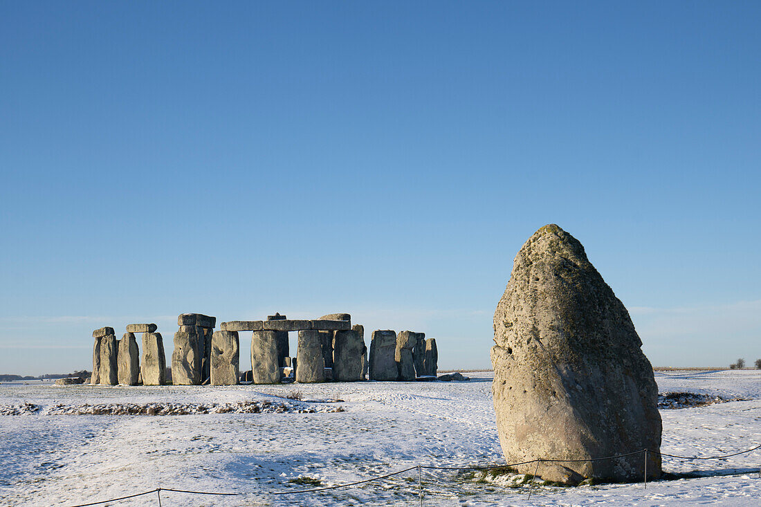 Stonehenge, geprägt von frühmorgendlichem Schnee mit blauem Himmel und dem Heel Stone rechts vom Steinkreis; Wiltshire, England, Vereinigtes Königreich