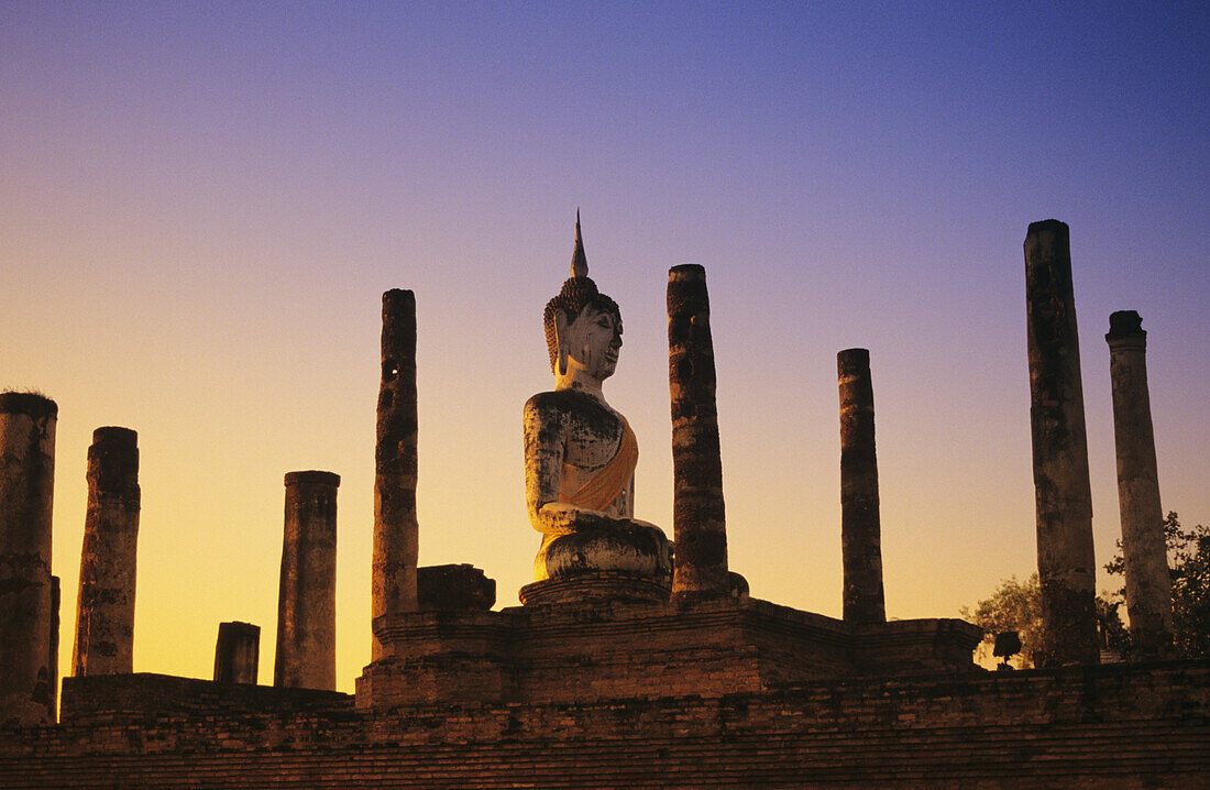 Thailand, Sukhothai, Wat Mahathat, Buddha-Statue mit vielen Säulen bei Sonnenuntergang, blauer und oranger Himmel.