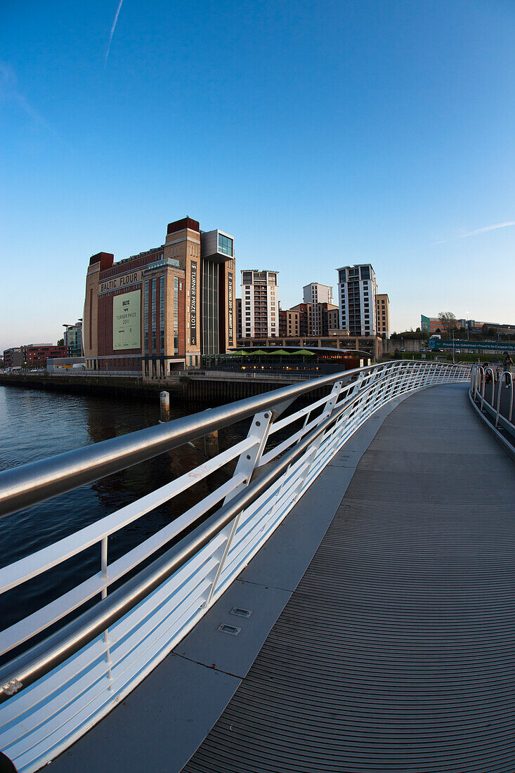 A Walking Bridge Over A River With Tall Buildings In The Distance; Gateshead Tyne And Wear England