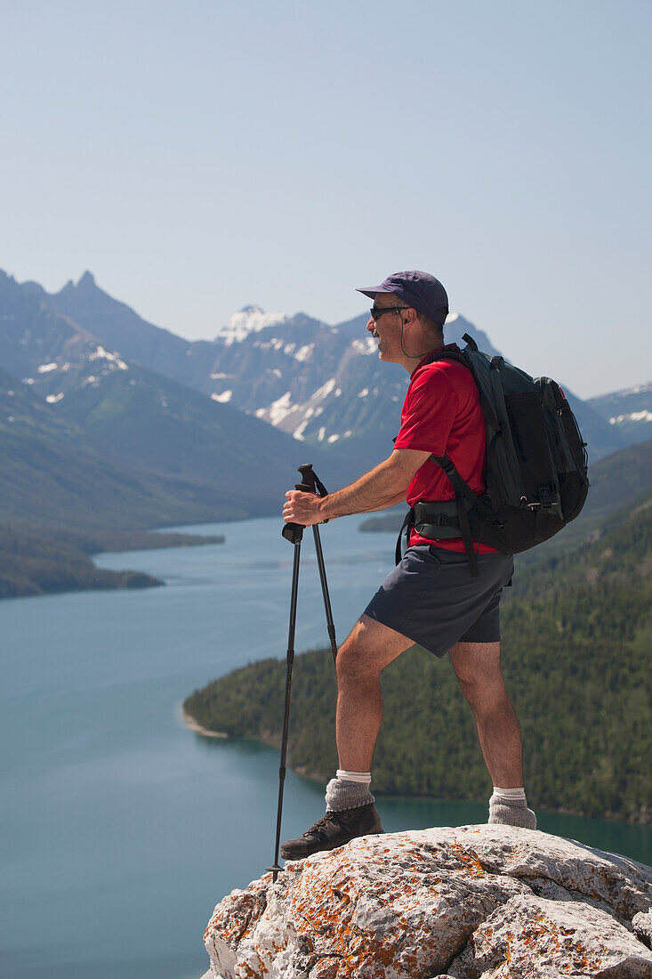 Male Hiker With Backpack And Hiking Poles Standing On A Rock Cliff Looking Out Onto A Lake With Mountains And Blue Sky; Waterton Alberta Canada