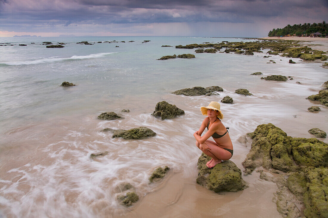 Eine Touristin hockt auf einem Felsen am Strand einer tropischen Insel; Koh Lanta Thailand.