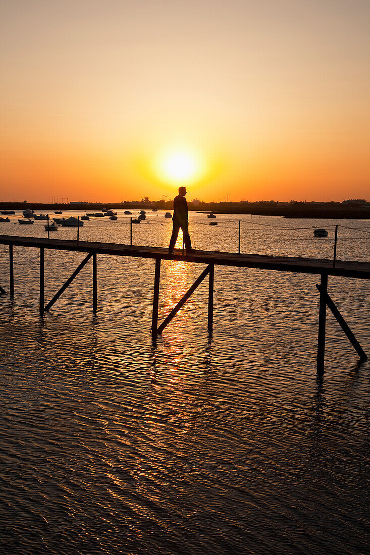 Ein Mann spaziert auf einem Pier mit im Wasser vertäuten Booten bei Sonnenuntergang in der Nähe von Faro; Algarve Portugal