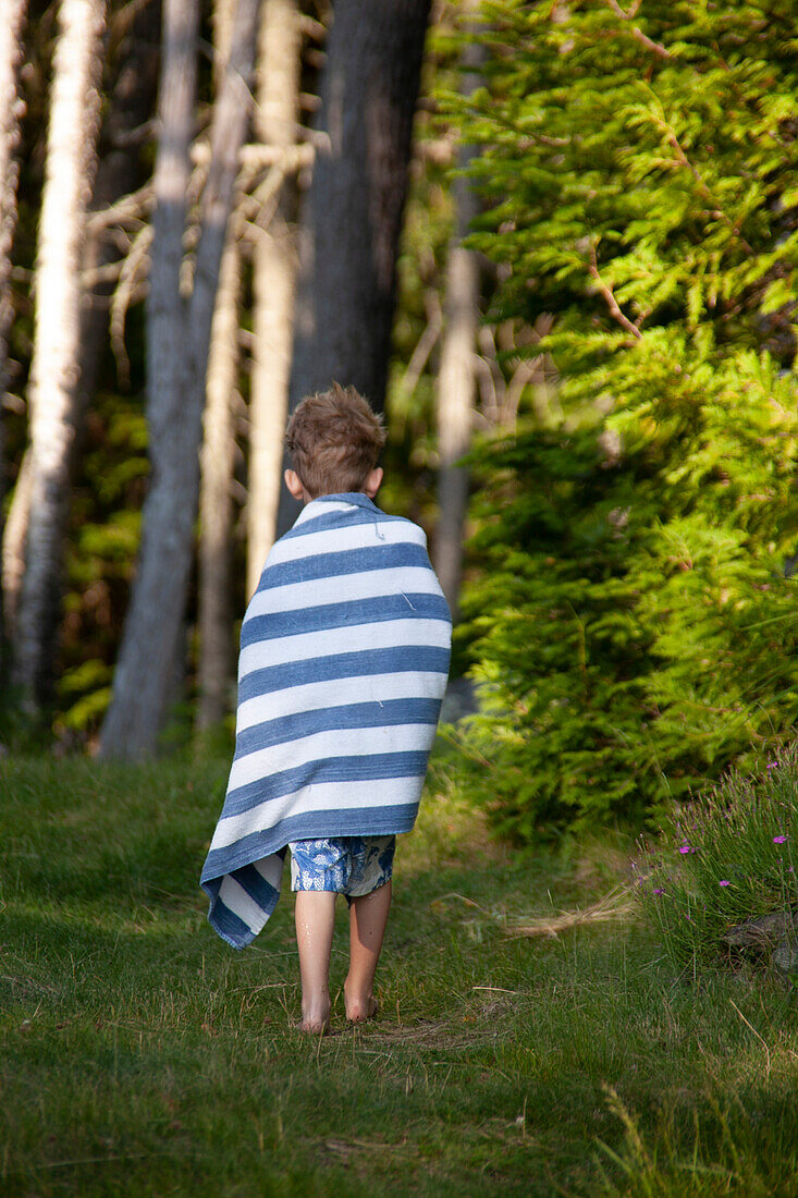 Rückansicht eines Jungen, der in ein gestreiftes Handtuch gewickelt einen grasbewachsenen Weg entlanggeht