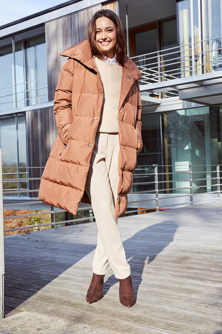 Young brunette woman wearing a beige sweater, matching trousers, and a brown quilted jacket