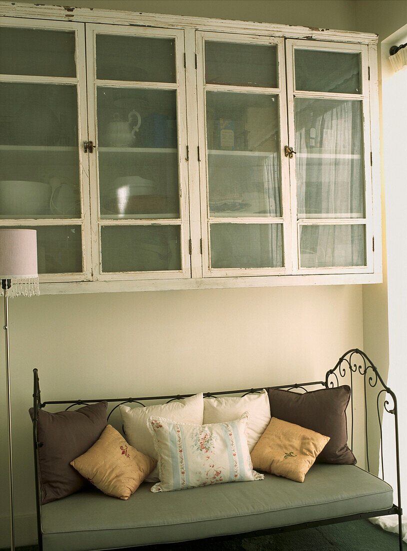 Glass fronted wall cabinets above upholstered wrought iron seat in kitchen