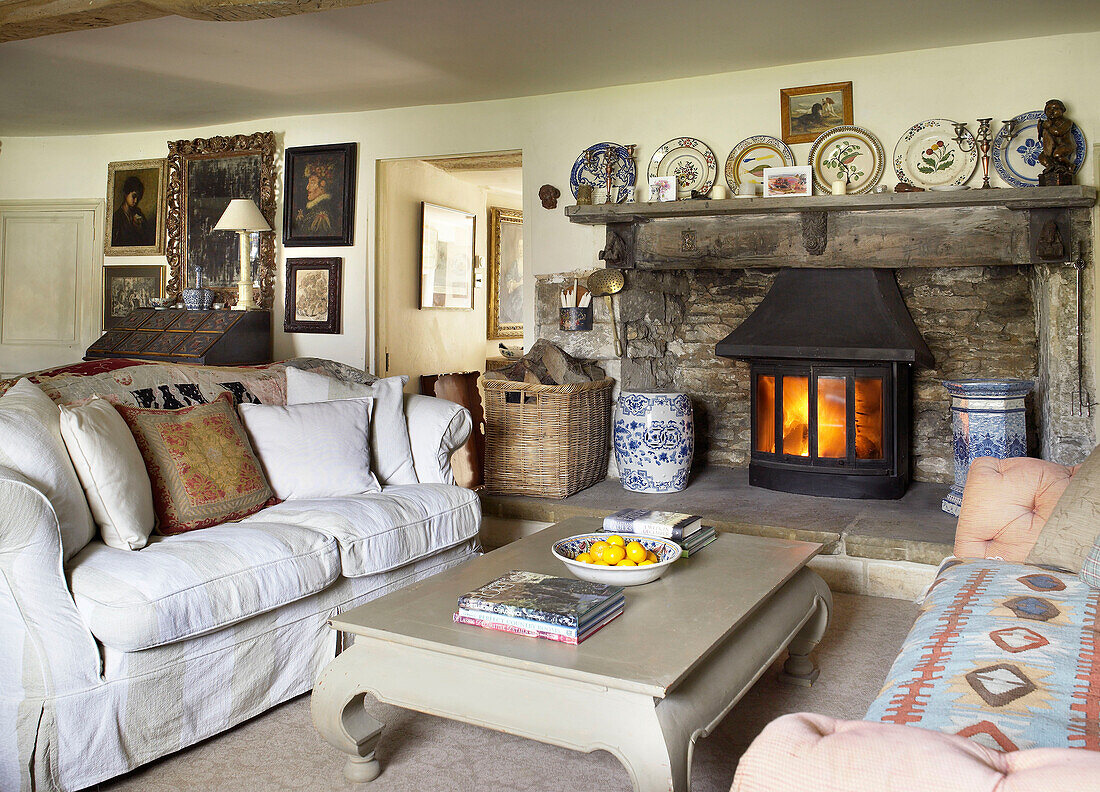 White sofa at fireplace with decorative chinaware in living room of Gloucestershire cottage, England, UK