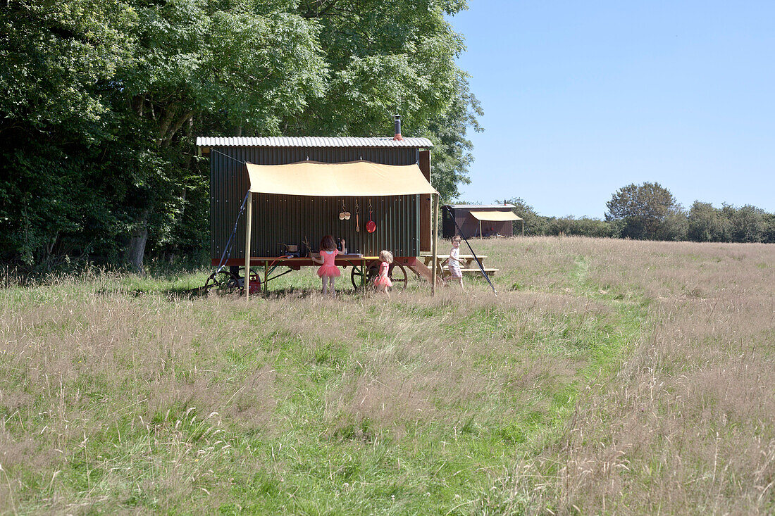 Three children beside Shepherds Hut in camping field of long grass