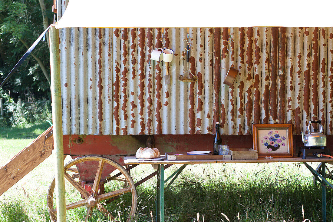 Outdoor kitchen are with trestle table under canvas canopy beside rustic Shepherds Hut in field