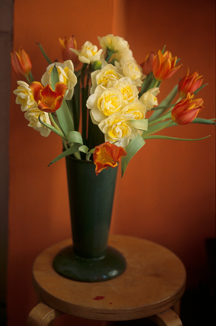 Frühlingsblumen in grün glasierter Vase auf einem Hocker aus Sperrholz