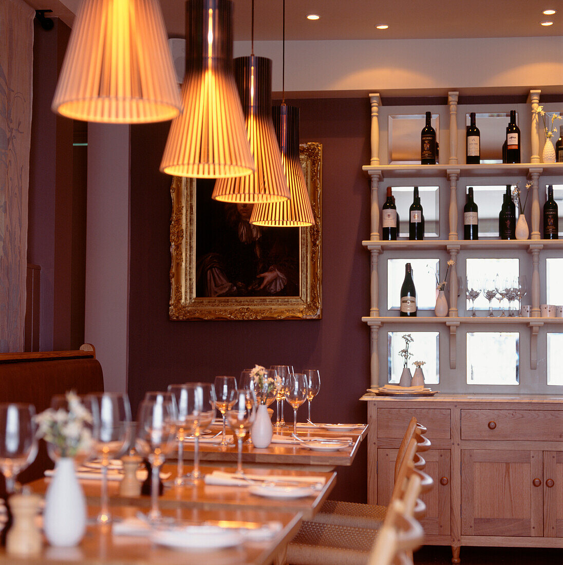 Restaurant table setting ready for guests with illuminated pendent lights and dresser with wine bottles