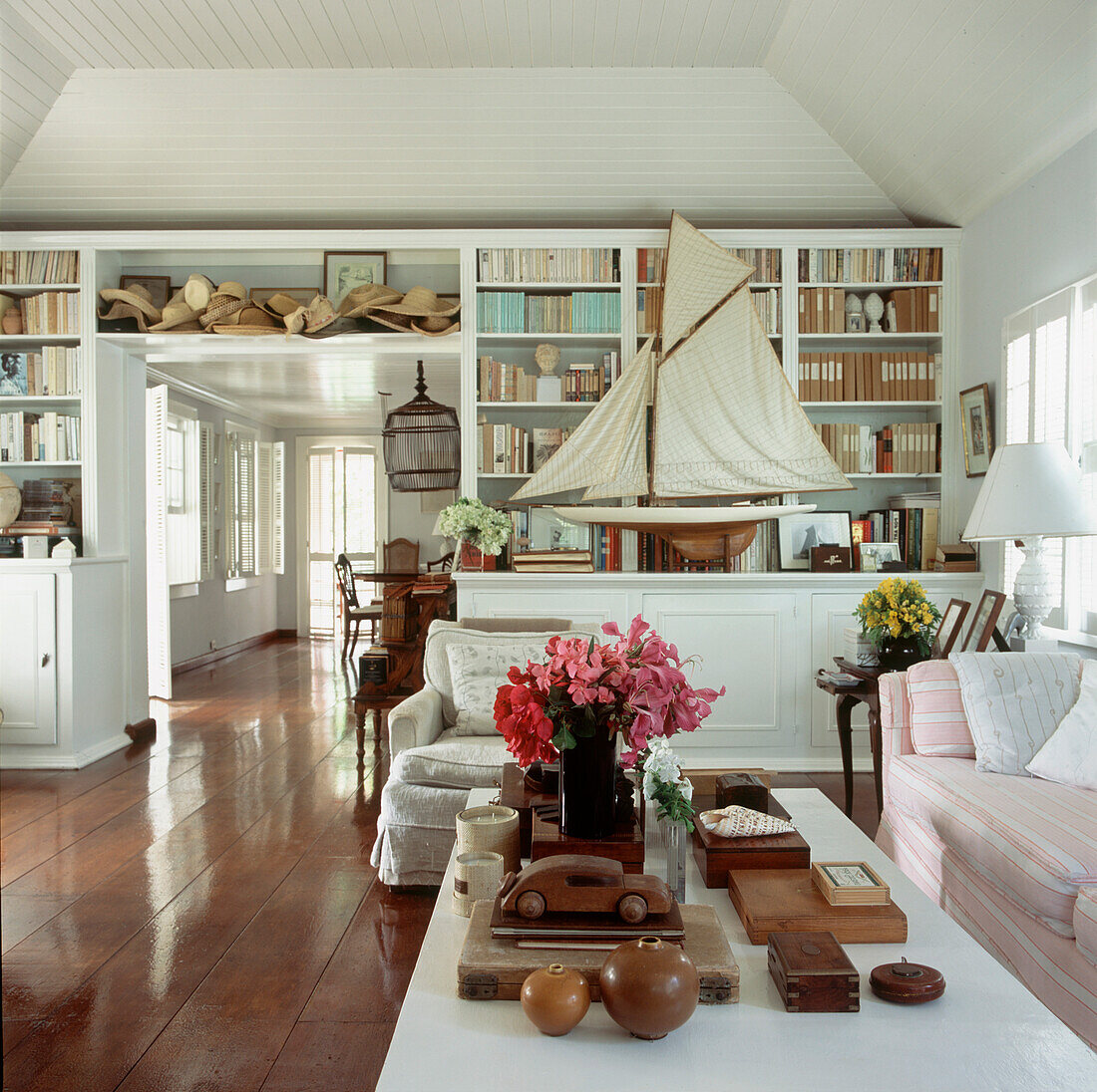 Open plan living dining room with built in bookshelves and cupboards displaying straw hats and a model sailing boat amongst other collectibles