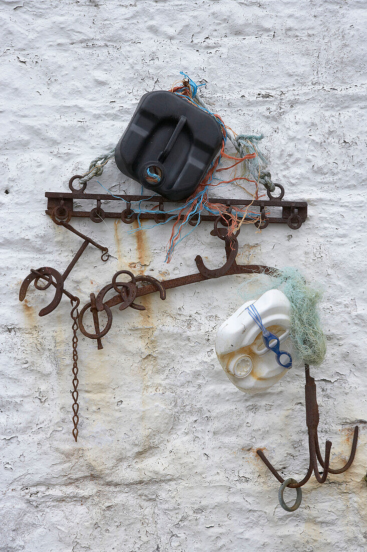 Weiße Außenwand mit geborgenen Strandfunden, darunter Plastikdosen und hängende Metallgegenstände