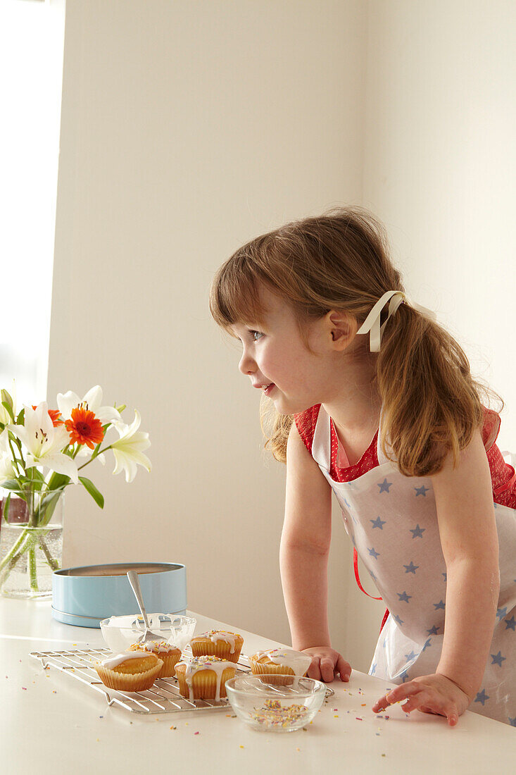 Drei Jahre altes Mädchen lehnt an einem Tisch mit einem Abkühlgitter und gefrorenen Fairy cakes