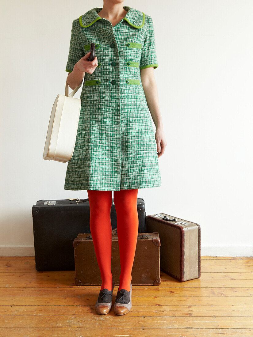 Frau in roter Strumpfhose und grünem Kleid steht mit alten Koffern