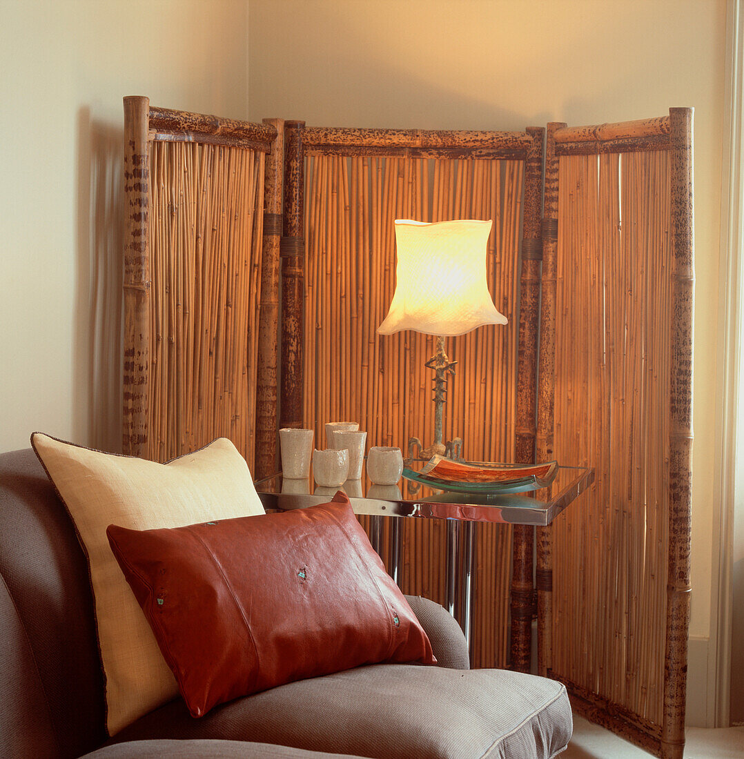 Room set with a bamboo screen behind a glass top table with table lamp and in the foreground a grey sofa