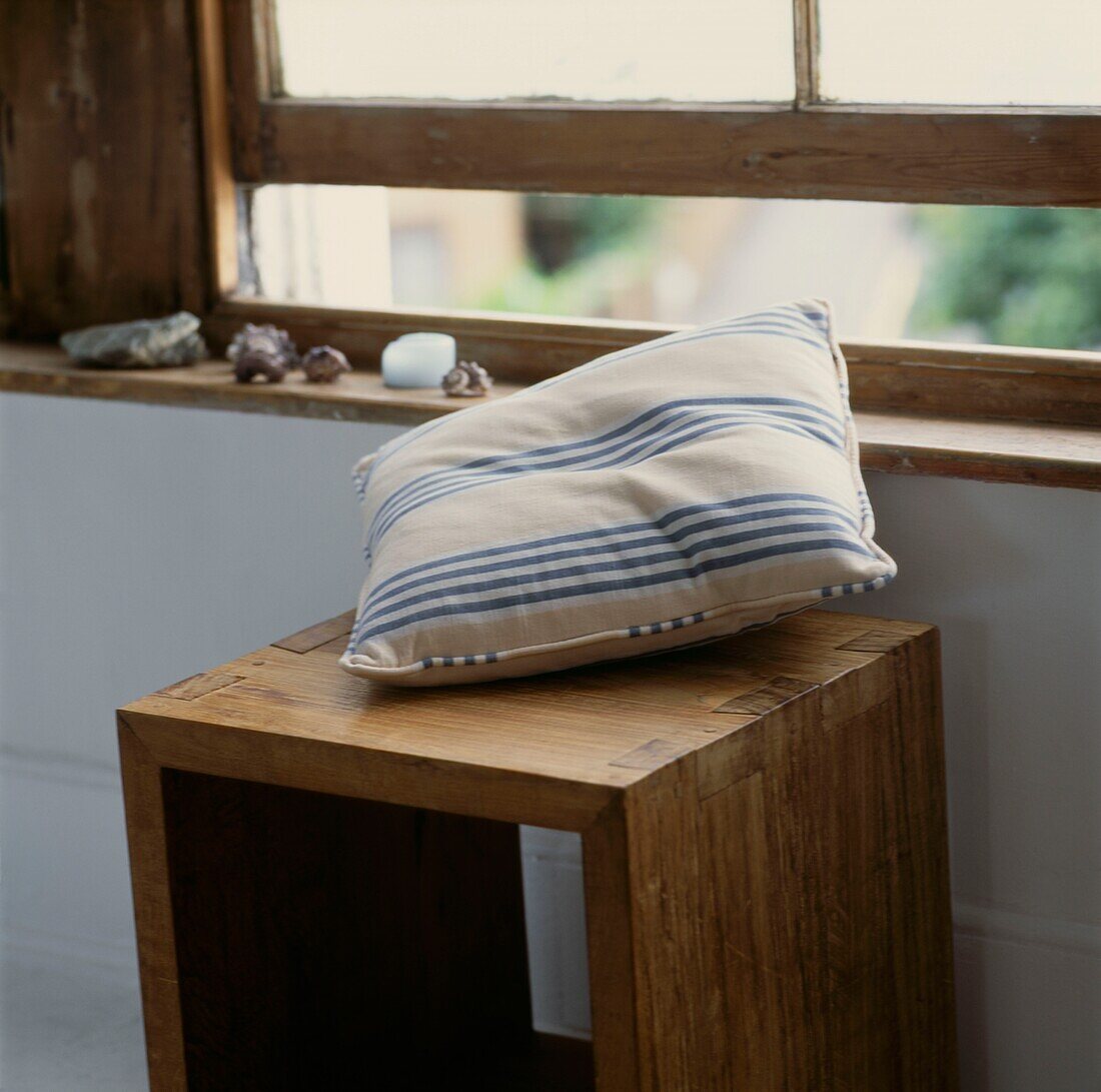 Gestreiftes Kissen auf einem Hocker an einem offenen Schiebefenster aus unbehandeltem Holz