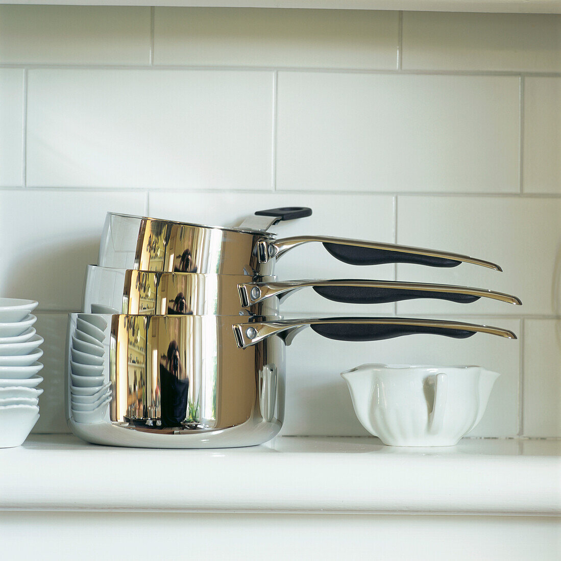 Shiny new stainless steel saucepans with white crockery on a shelf