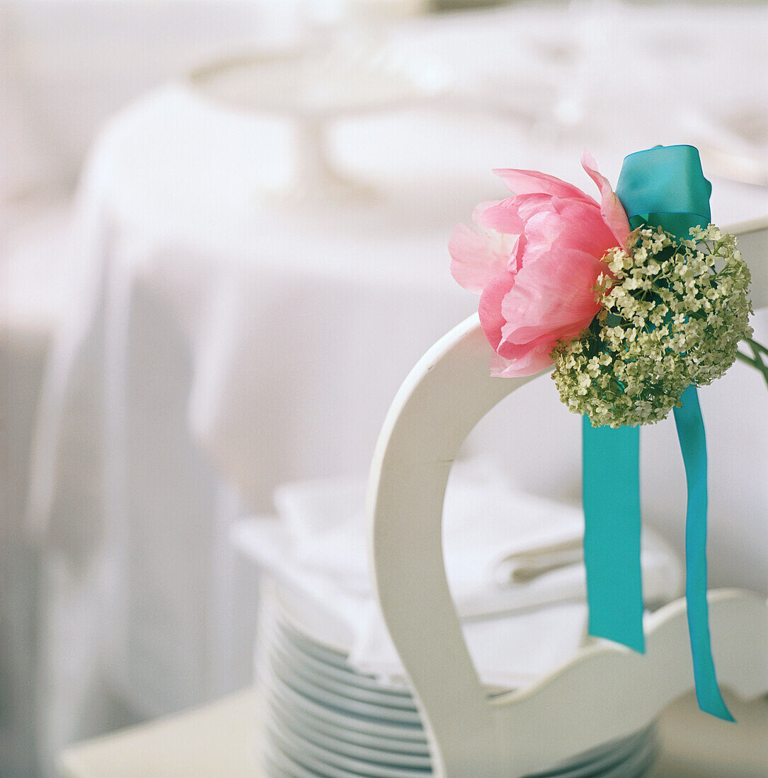 Back of white chair tied with a bouquet of flowers and ribbon in a reception area with plates and napkins stacked on the chair