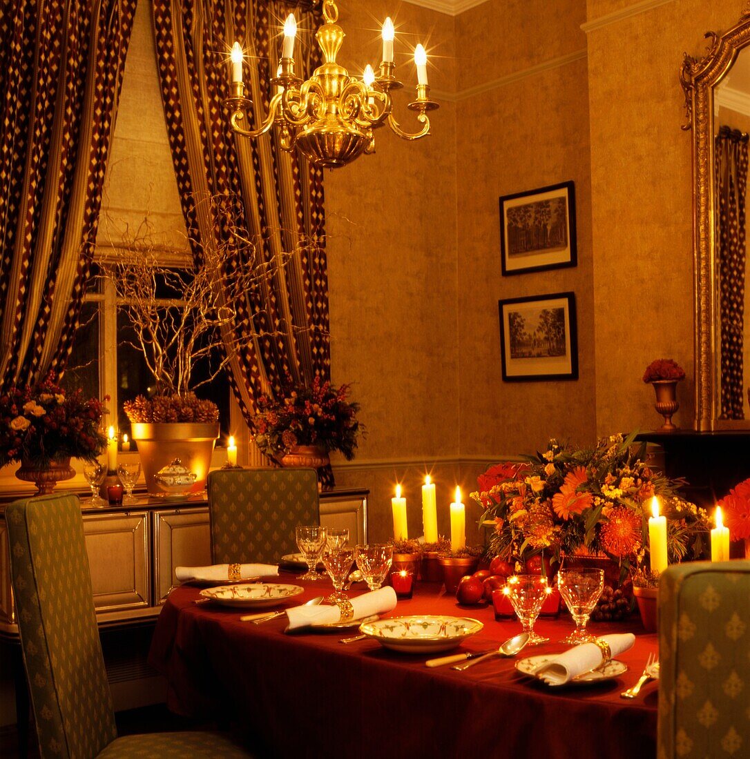 Lit candles on Christmas dinner table with gold chandelier and tied back patterned curtains 