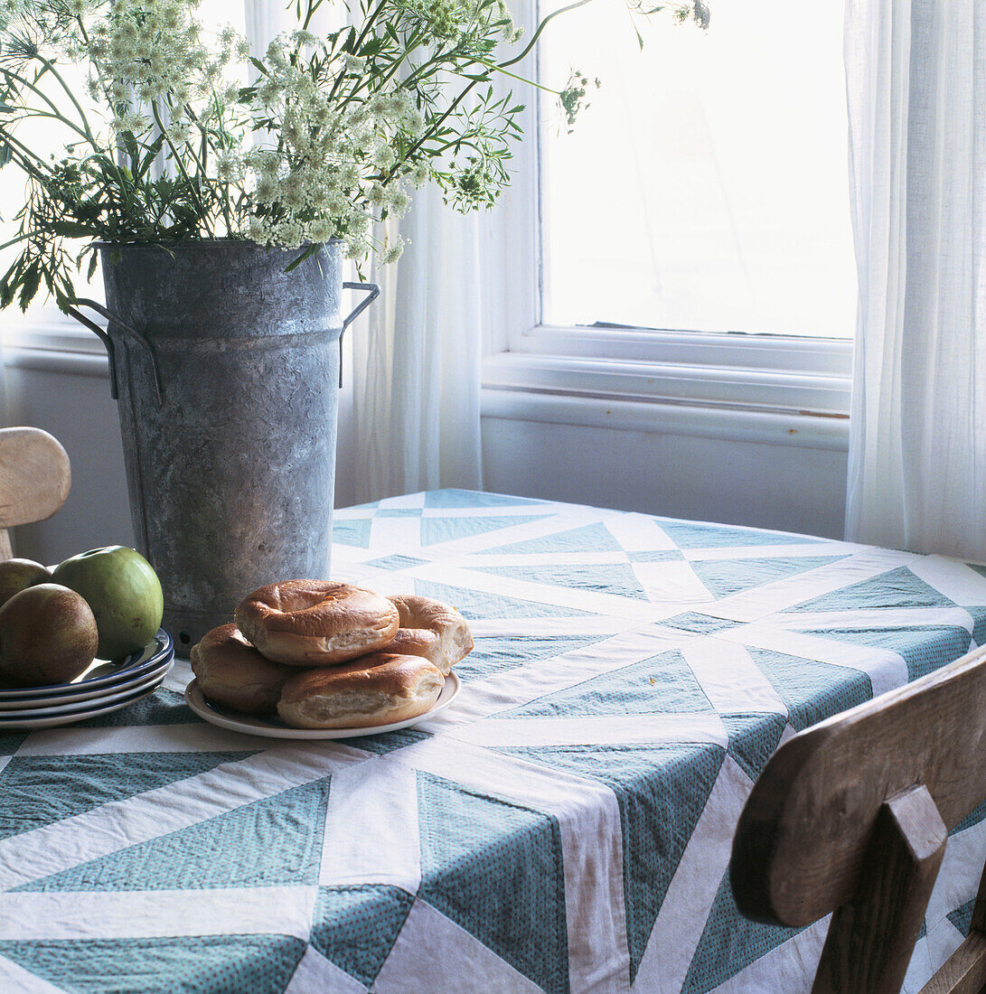 Bagels, Äpfel und Wildblumen auf einer Patchwork-Tischdecke neben einem Fenster in der Küche
