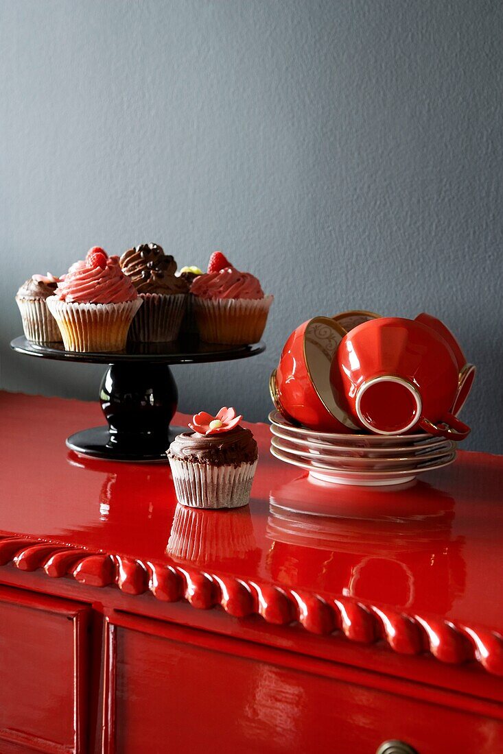 Bright Red painted sideboard against a grey wall with red cups and saucers stacked beside a cakestand filled with cup cakes