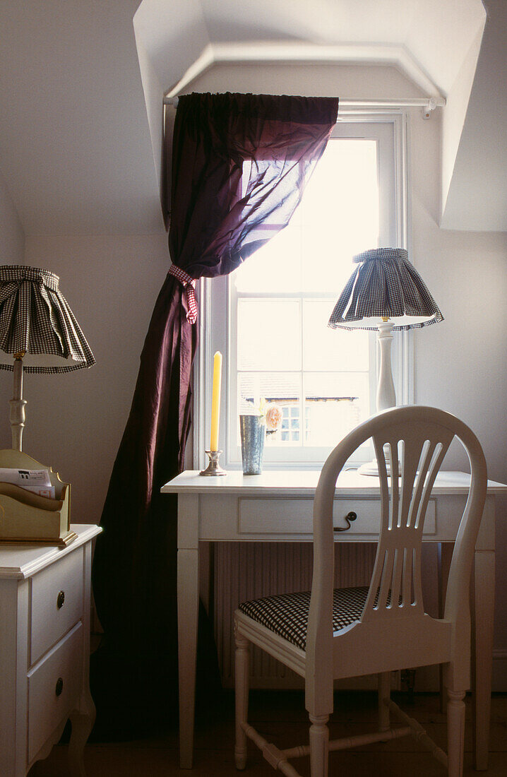 Desk and lamp in window recess dressed with dark red unlined silk drapery
