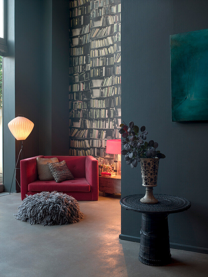 Pink armchair beside a wall decorated in bookcase wallpaper with grey floor cushion and side table with vase