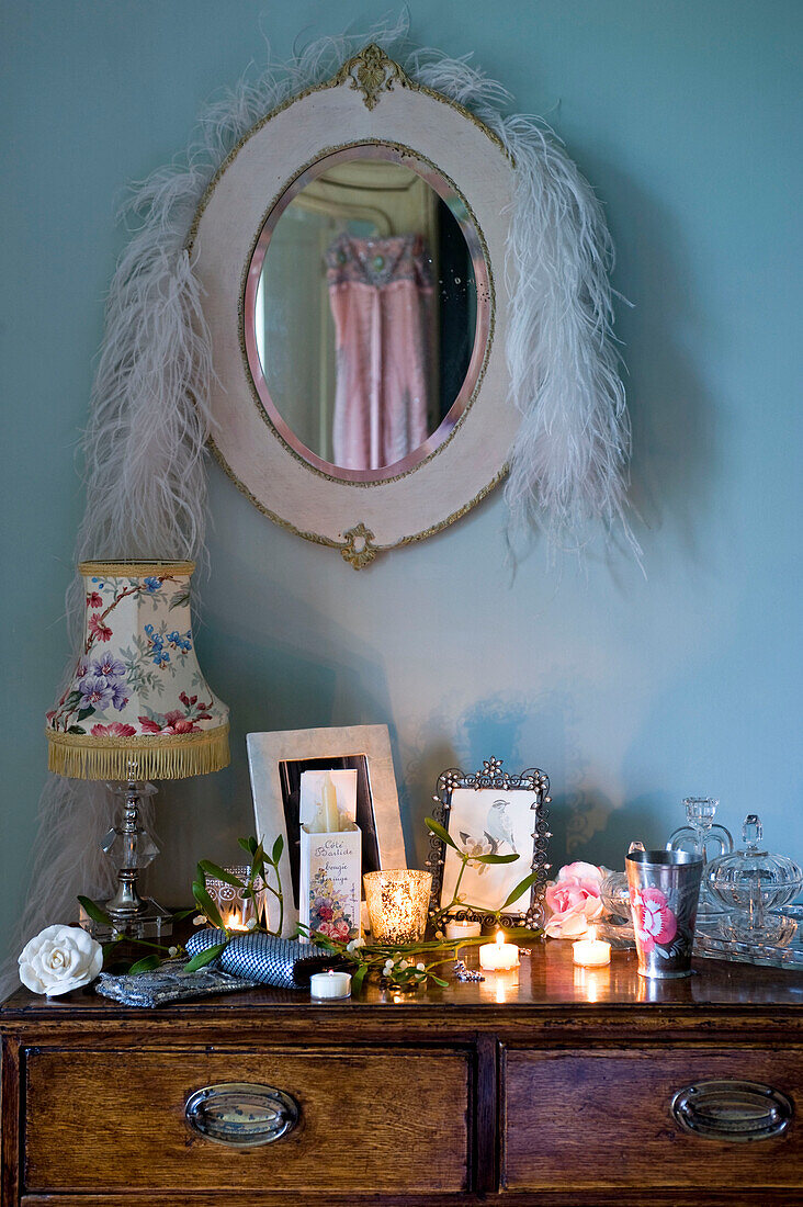 White feather boa hangs over mirror above wooden side unit with picture frames and lit candles