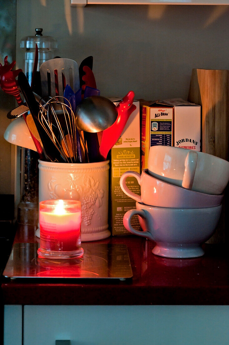 Utensil holder and lit candle with stacked teacups in kitchen of London home UK
