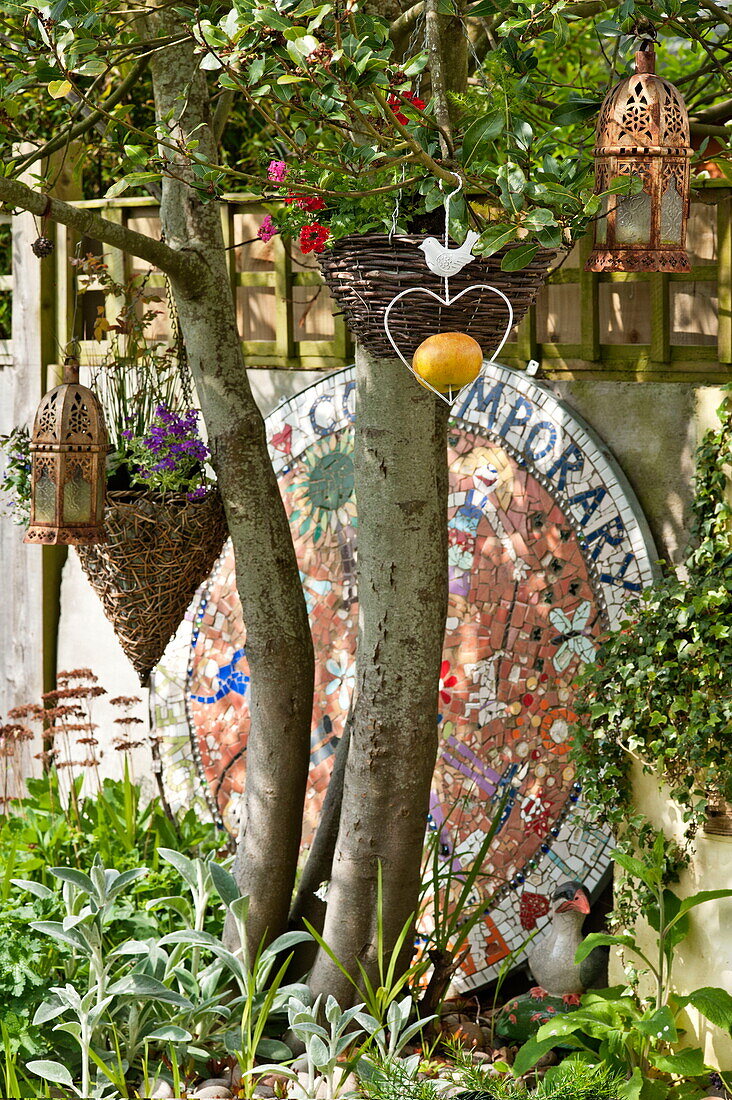 Garden ornaments hang from tree with large mosaic in garden of Bovey Tracey, Devon, England, UK
