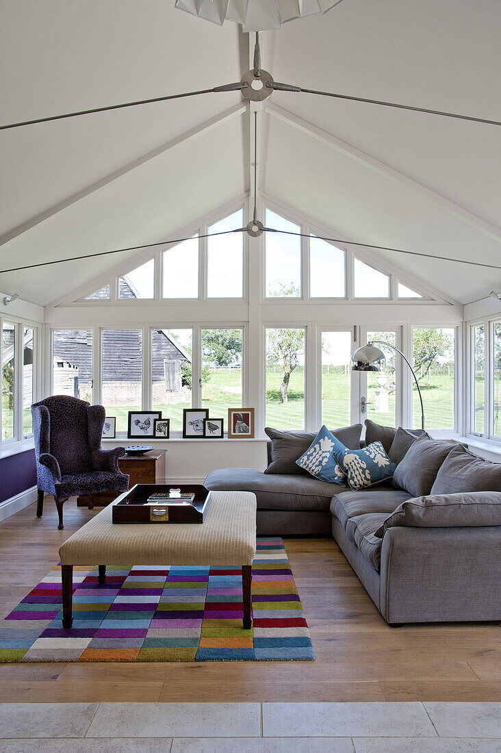 Graues Sofa mit buntem Teppich und Hocker im Wintergarten eines modernen Landhauses in Suffolk, England, UK