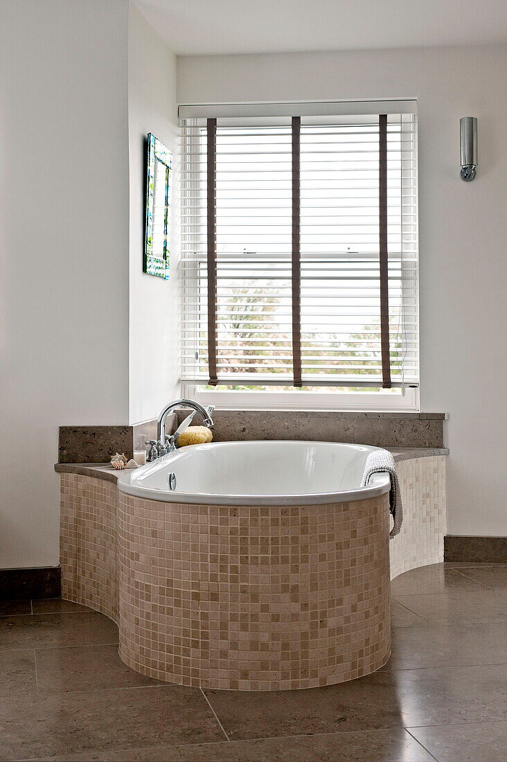 Mosaic tiled bath at window with Venetian blinds in contemporary home, Cornwall, England, UK