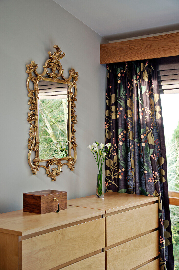 Gilt framed mirror above chest of drawers with floral curtains in Cornwall bedroom, UK