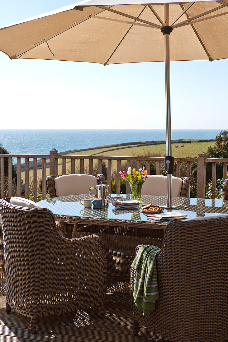 Wicker chairs at table with parasol on terrace, Cornwall, UK