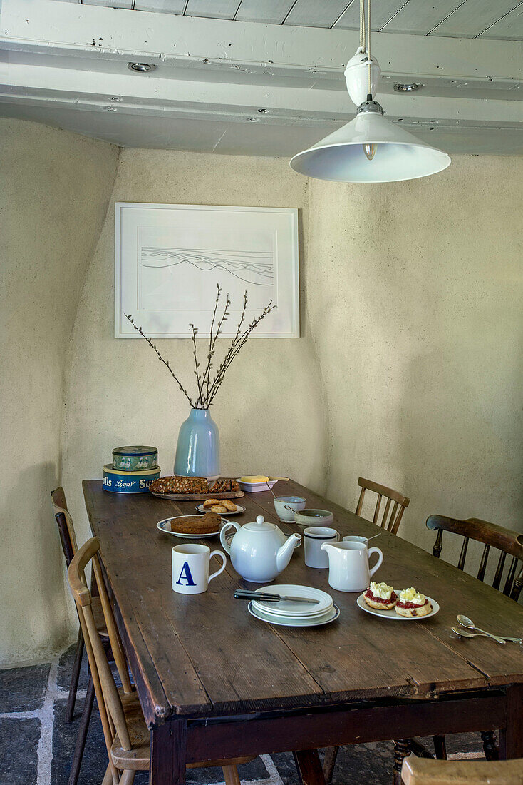 Holztisch unter weißer Hängelampe mit Kunstwerk und Scones für den Nachmittagstee in einem Bauernhaus in Helston, Cornwall, UK