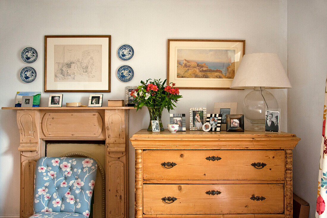 Familienfotos auf einer hölzernen Kommode mit einem blumengemusterten Stuhl am Kamin in einem Cottage-Schlafzimmer in Cornwall, Großbritannien