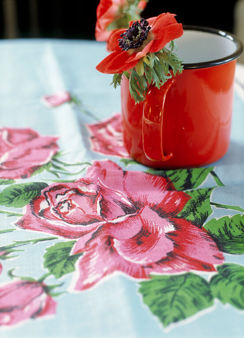 Rote Anenome in Emaille-Tasse auf Tischtuch mit Blumenmuster