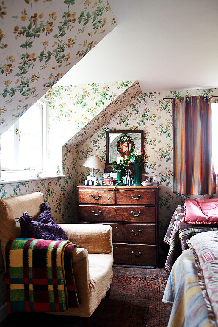 Armchair and wooden chest of drawers below dormer window in floral papered bedroom of Shropshire cottage England UK