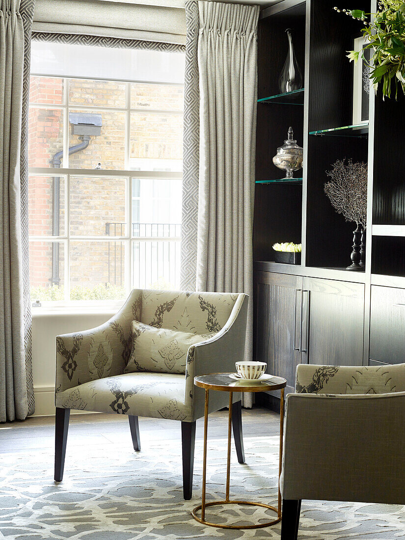 Pair of armchairs with brass side table and storage shelves at window in London townhouse, UK