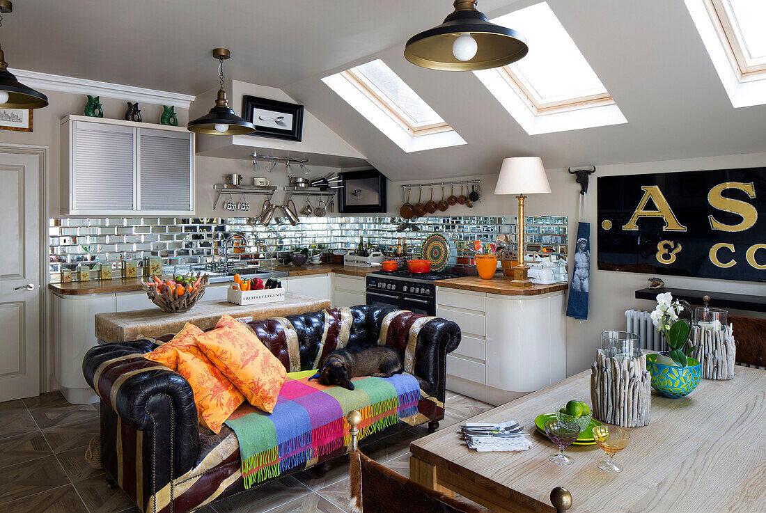 Chesterfield sofa under skylight windows in open plan attic kitchen with mirrored splashback in London home, England, UK