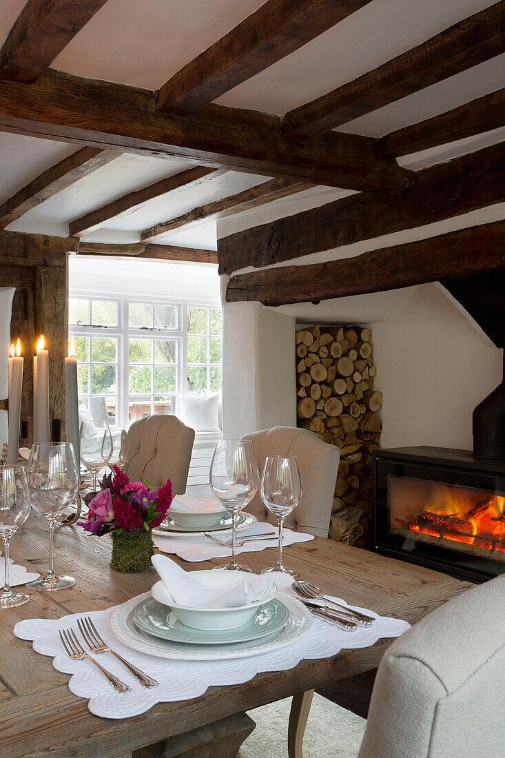 Beleuchtetes Feuer und Kerzen mit Gedeck auf einem hölzernen Esstisch in einem Haus in Surrey, England UK