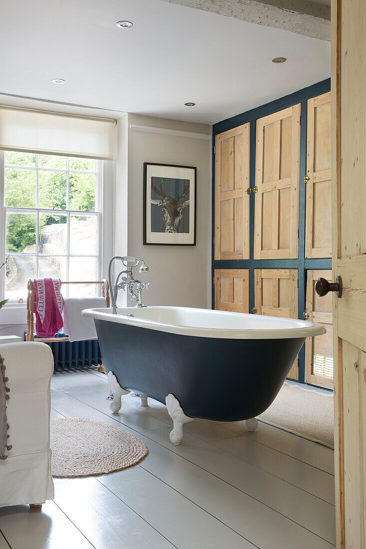 Dark blue freestanding bath with wood panelled cupboards in Gloucestershire home England UK